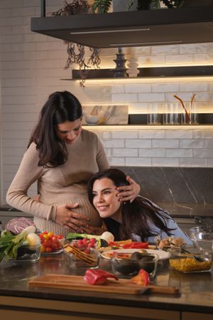 Pregnant woman helping friend to hear heartbeat of baby in kitch
