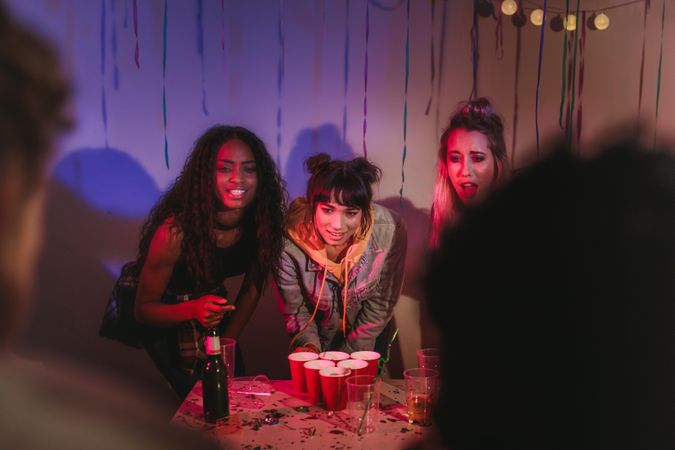 Three young women standing at a table with glasses and a bottle arranged for the house party