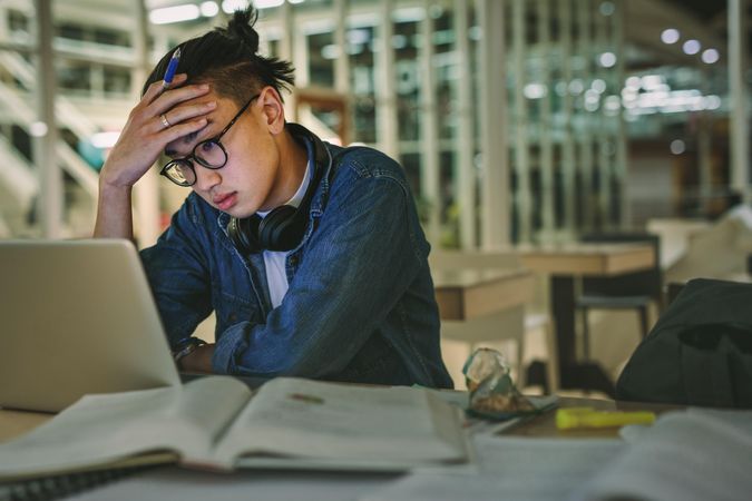 Male student sitting at library looking at laptop with hand on forehead