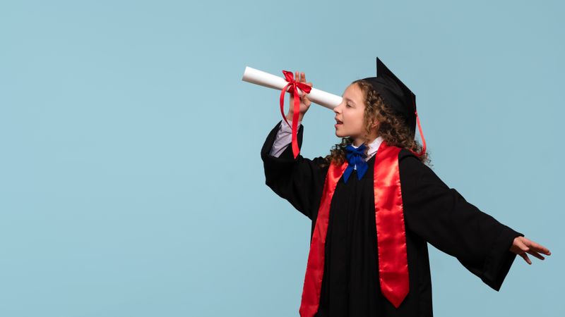 Young girl in graduation gown holding diploma like telescope.
