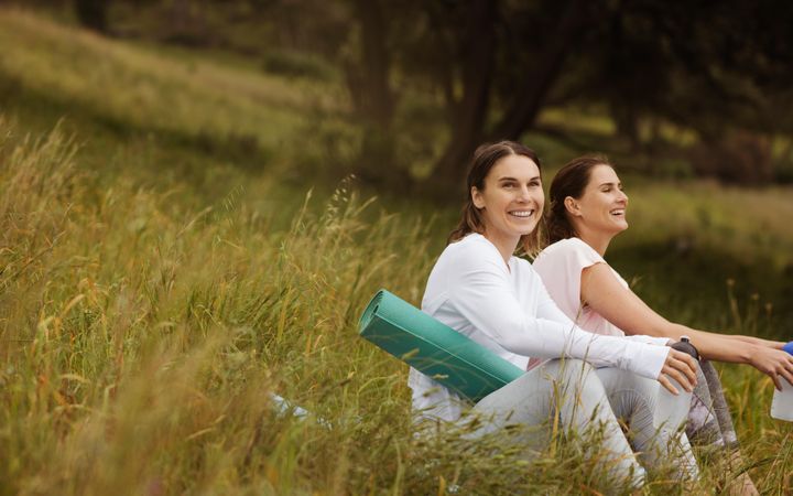 Two cheerful women friends sitting with yoga mats in the meadows