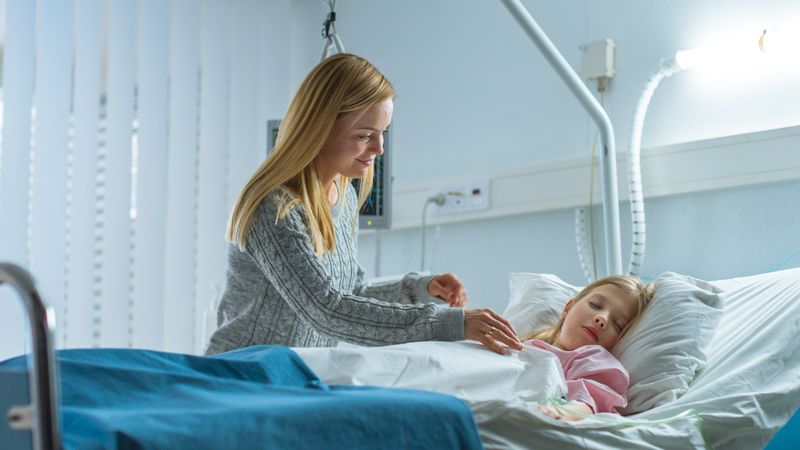 Cute Little Girl Sleeps on a Bed in the Children's Hospital, Caring Mother Covers Her with a Blanket and Caresses Her Forehead