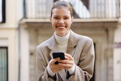 A joyful young woman is smiling brightly while she is texting on her smartphone outdoors