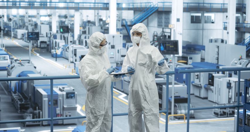 Female Workers Wearing Full-Body Polypropylene Coveralls at a Manufacturing Plant. Specialists Using Tablet Computer, Discussing Automated Work Process. Employees Protected From Hazardous Materials