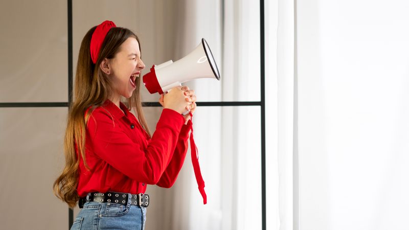 Teen girl shouting into megaphone near bright window.