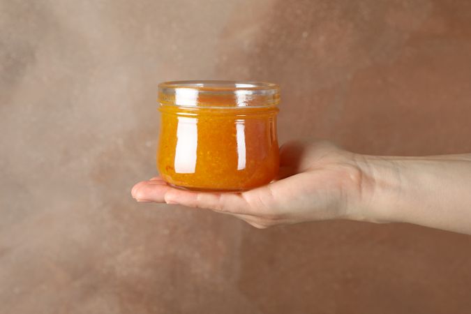 Female hand holds glass jar with apricot jam on brown background