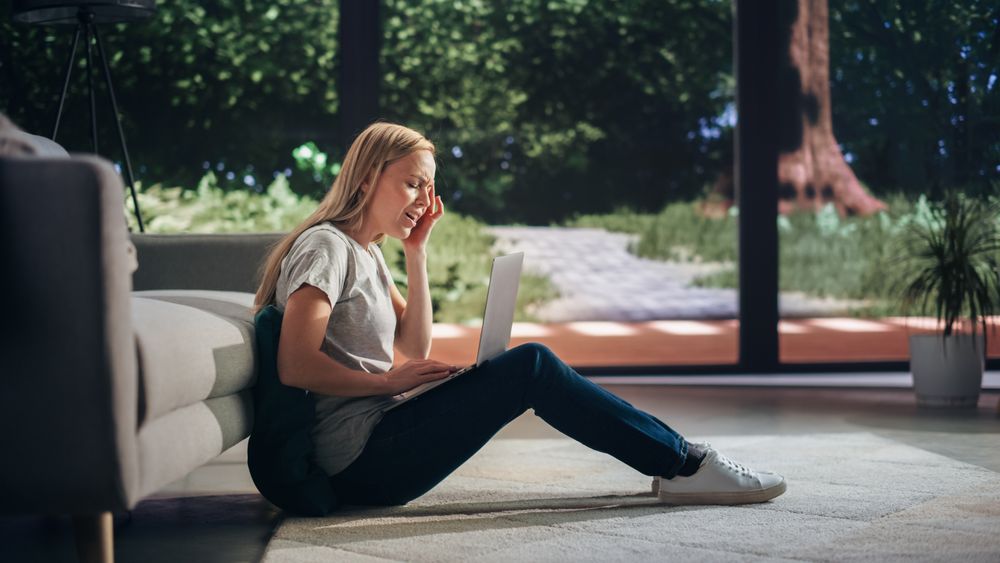 Portrait of a Young Adult Female Experiencing Sudden Migraine while Sitting on a Floor and Using Laptop Computer in Sunny Living Room.
