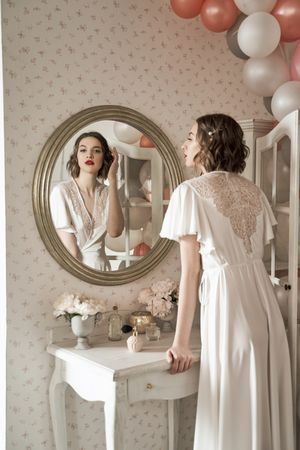 serious young woman standing and looking in big round mirror in boudoir
