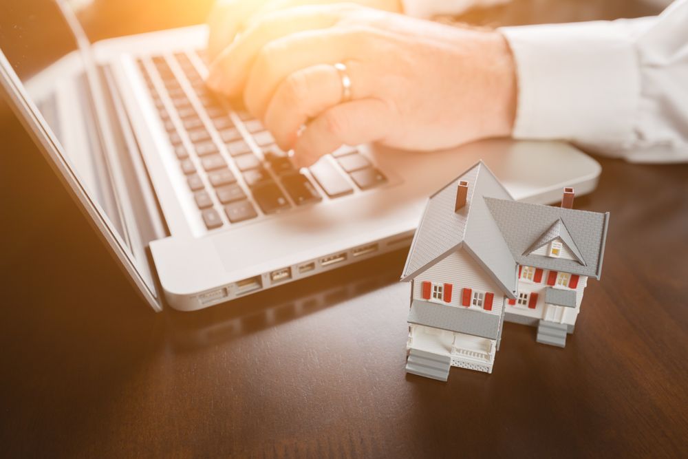 Miniature House Resting on Desktop And Man Using Laptop Computer.