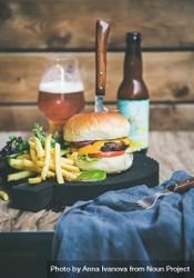 Classic hamburger skewered with knife with fries and beer bottle at wooden restaurant table 56NjV5