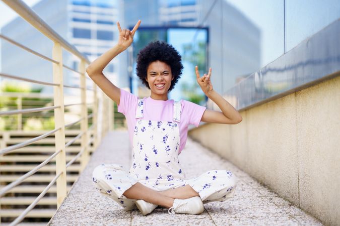 Woman in floral overalls sitting cross legged near hand rails with hands up