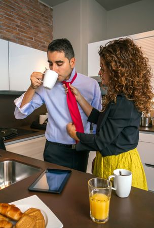 Curly haired woman adjusting tie of man while having fast breakfast before work