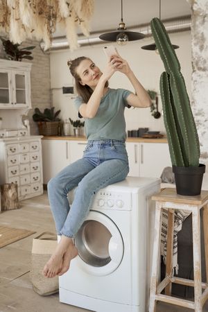 Relaxed women sitting on washing machine and browsing smartphone