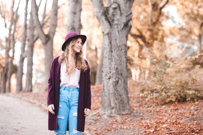 Smiling teenage girl in purple coat with hat standing near fallen yellow tree leaves