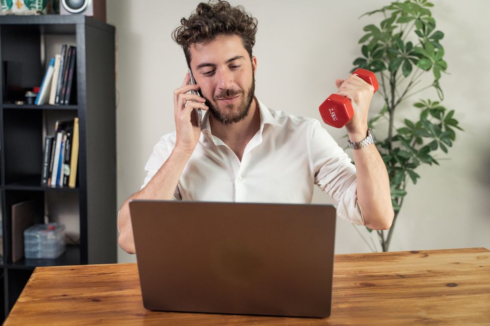 Young man multitasking with phone laptop and dumbbell in modern workspace