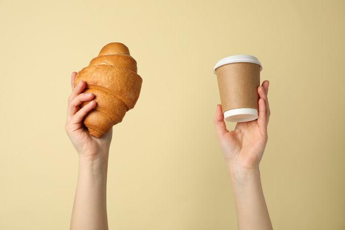 Female hands holding croissant and cup of coffee on beige background