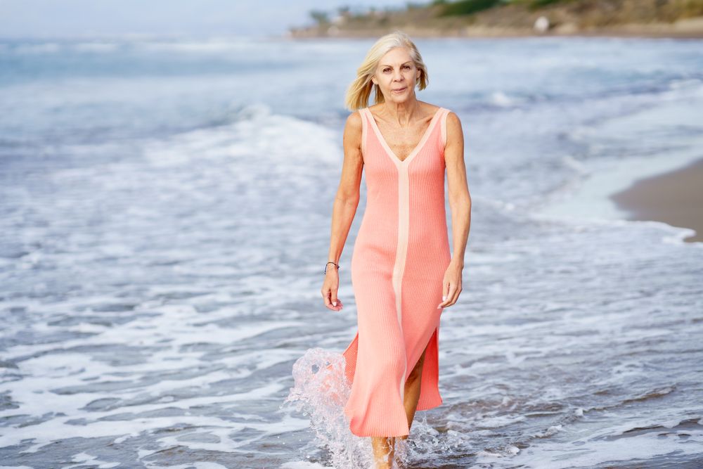 Mature woman in pink dress strolling along coast on beach