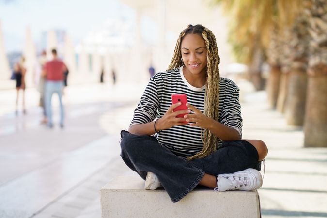 Smiling woman checking mobile phone while sitting in pedestrian area outside