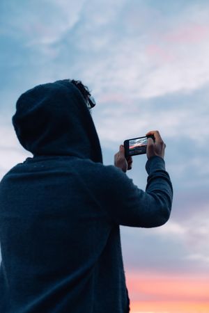Man wearing hood on cliff taking photo on phone at sunset