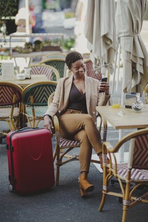 Smiling Black woman with smartphone sitting at table near luggage in cafe
