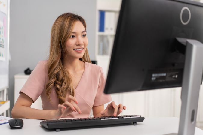 Happy Asian woman sitting in office and typing on keyboard while looking at monitor