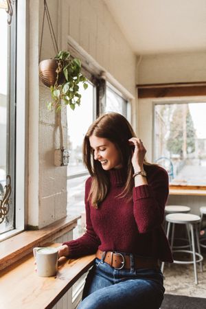 Woman in burgundy sweater sitting by the window holding mug