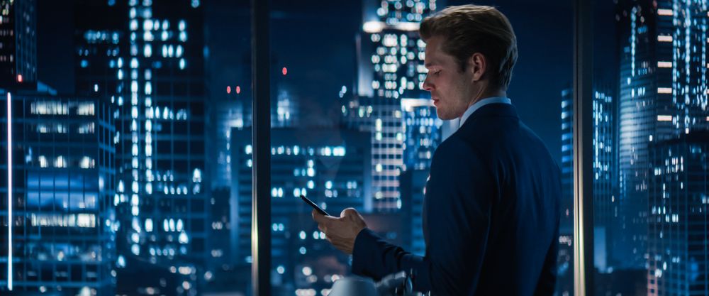 Sideview of Businessman in Stylish Suit Standing Next to Window in Big City Office with Skyscrapers Late At Night.