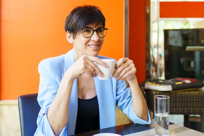 Positive female in trendy blue jacket and eyeglasses sitting at table with cup of hot drink and looking away