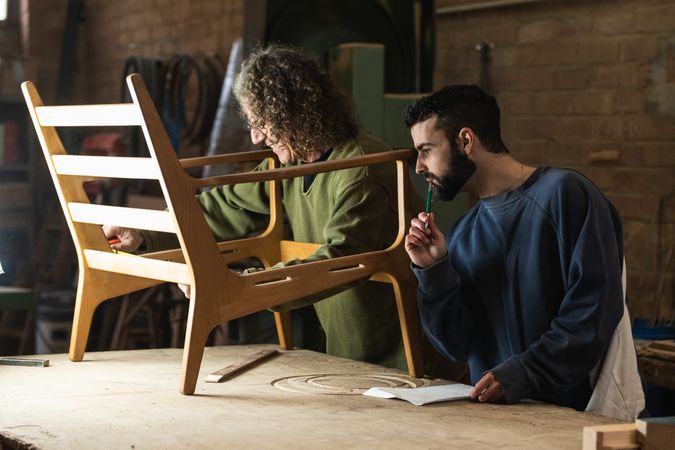 Craftsman creating wooden chair with coworker drawing sketches