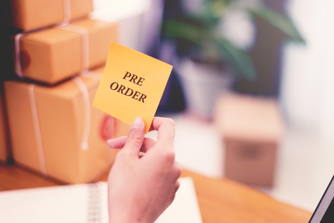 Hand holding post it note in front of boxes with the words “pre-order”