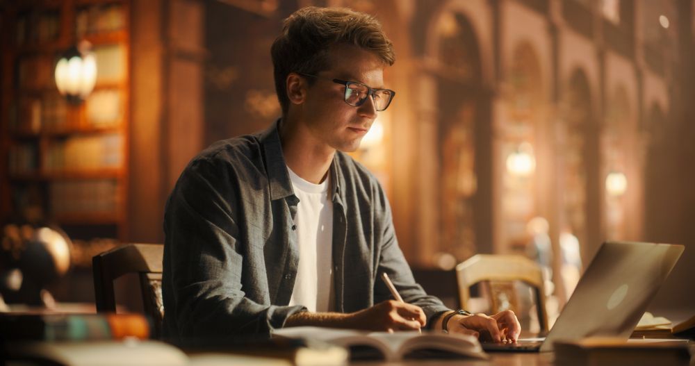 Concentrated Young Male Student Engaged in Academic Research With Laptop and Books in a Classical Library Setting.