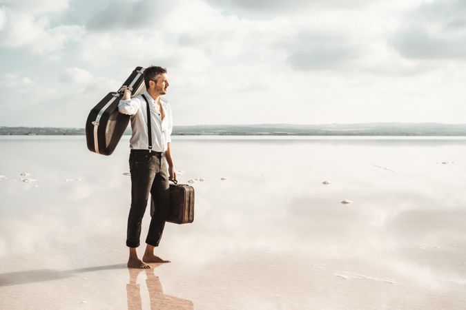 Focused stylish man with suitcase and guitar gig bag on seaside