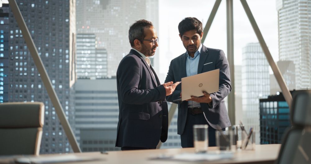 Two Young South Asian Businessmen Standing Next to a Window in a Modern Office with a City Skyline View.
