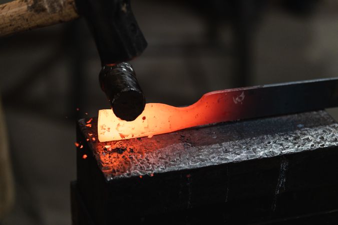 Blacksmith in gloves hot hitting metal part on anvil