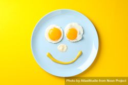 Looking down at blue plate on yellow table with smiley face on it made of eggs and condiments 5wBPW0