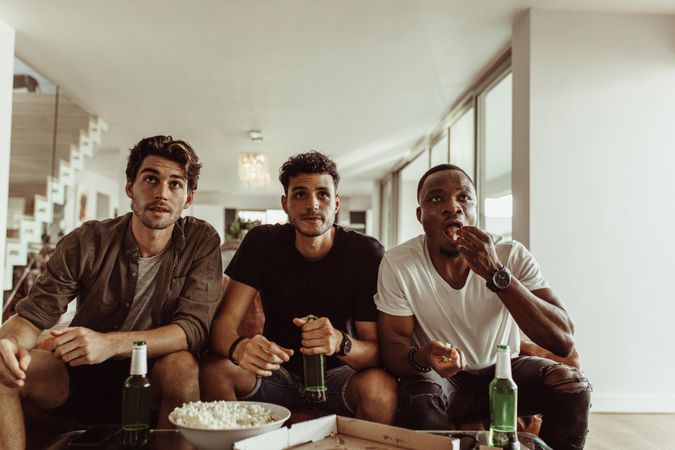 Men sitting on sofa with drinks and snacks watching television at home