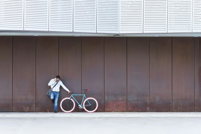 Male in sunglasses standing with red and green bicycle in front of wall and speaking on phone