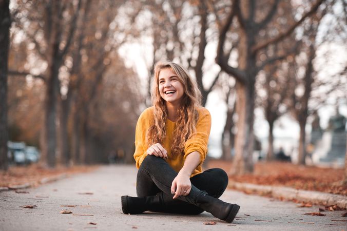 Smiling teen girl in yellow sweater sitting on pathway near bare trees