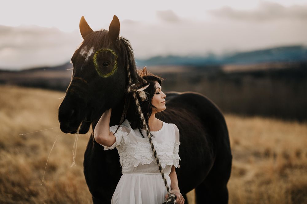 Woman in light dress standing beside dark horse in yellow grass field