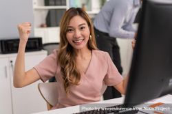 Asian woman making fists with her arms up in celebration while sitting at her office desk 4O6Ga5