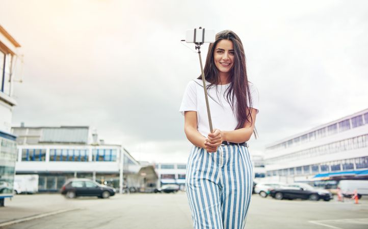 Brunette woman in striped shirt taking a picture with a selfie stick outside