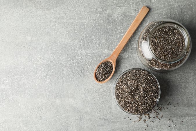 Glass bowl, jar and wood spoon with chia on grey background, copy space