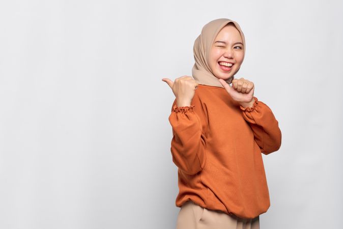 Smiling Muslim woman in headscarf and orange sweater gesturing behind her with thumb and winking