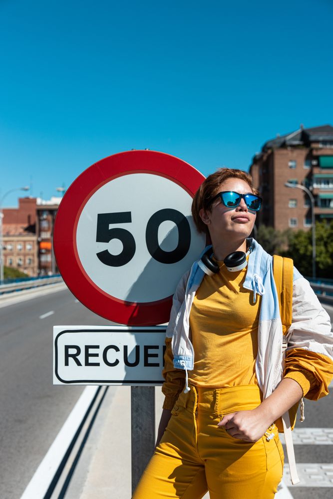 Trendy joyful young woman in sunglasses enjoying view beside road sign on bridge in sunny day