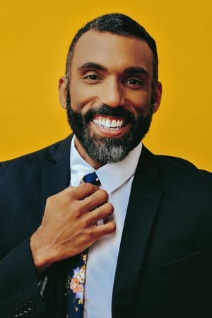 Happy Black male in suit fixing his floral tie in yellow studio