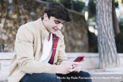 Young man using mobile phone and smiling while sitting outdoors bE997M