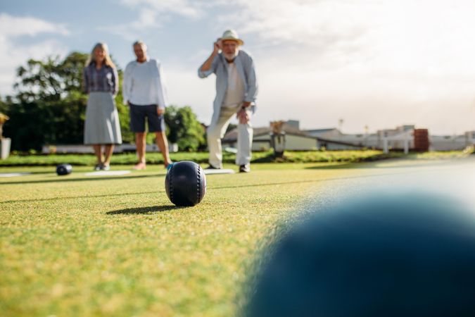 Older man in hat throws a boules standing in position in a lawn with friend standing behind