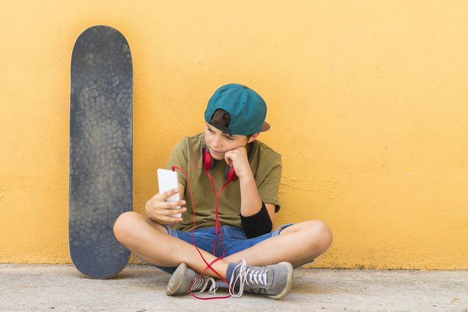 Teenage boy sitting on ground leaning on a yellow wall and checking phone