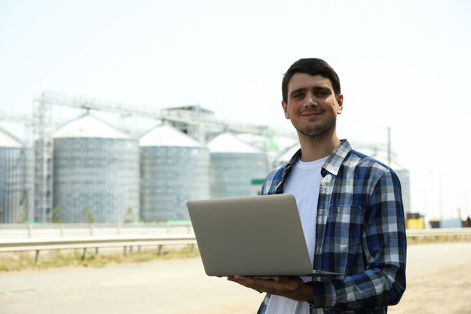 Young man with laptop against grain silos. Agriculture business