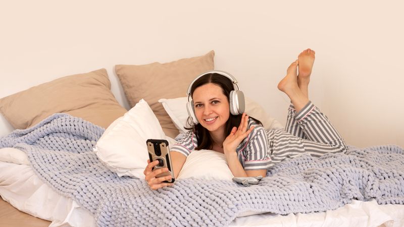Woman in bed with headphones, smiling and waving.